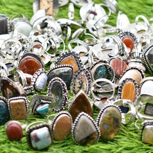 Op de afbeelding: Een verzameling zilveren ringen met verschillende gekleurde edelstenen. De ringen zijn gerangschikt op een groene grasmat.