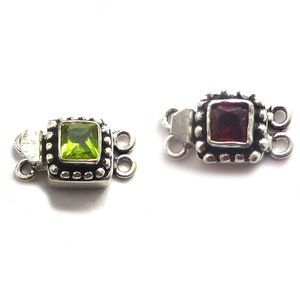 Op de afbeelding: Een set van tien zilverkleurige sieraden sluitingen met verschillende gekleurde edelstenen. De sluitingen zijn vierkant met een decoratieve rand en hebben een enkele edelsteen in het midden. De edelstenen zijn paars, blauw, groen, rood en geel.