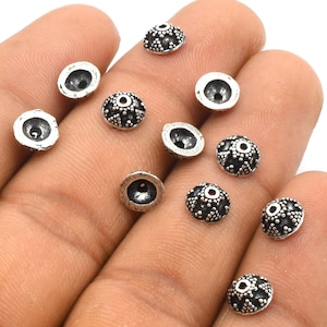 May include: A close-up of ten silver-toned metal bead caps with a decorative design. The bead caps are round and have a small hole in the centre.