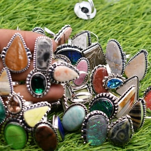 May include: A collection of silver rings with various gemstone inlays, including green, blue, red, and brown. The rings are displayed on a green grassy surface.