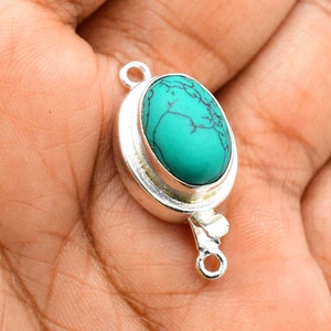 Puede incluir: Un colgante ovalado plateado con una piedra turquesa. El colgante tiene un bucle en cada extremo para sujetarlo a una cadena o cordón.