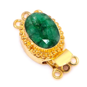 Pode incluir: Um fecho de joia dourado com uma pedra preciosa de esmeralda oval e facetada no centro. O fecho tem uma borda decorativa com contas ao redor da esmeralda e uma barra retangular com três laços.