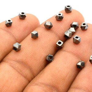 Puede incluir: Una colección de pequeñas cuentas de metal negro facetadas. Las cuentas están dispuestas en una mano y parecen estar hechas de un metal brillante.