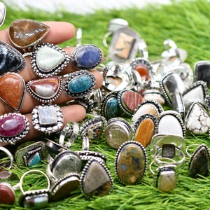 Puede incluir: Una colección de anillos de plata con diferentes incrustaciones de piedras preciosas. Los anillos presentan una variedad de formas y tamaños, incluyendo ovalados, redondos y en forma de lágrima. Las piedras preciosas son una mezcla de colores, incluyendo rojo, azul, verde, marrón y blanco.