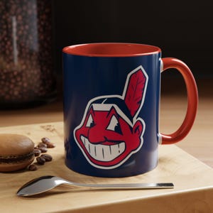 May include: A navy blue ceramic mug with a bright red interior and handle. The exterior features a large, graphic illustration of a smiling cartoon character with a red face, white teeth, and a single red feather in a black headband centred on the mug.