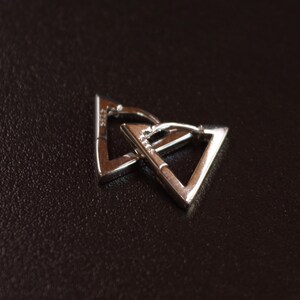 May include: A pair of silver triangle-shaped hoop earrings. The earrings are interlocked, creating a unique geometric design. The jewellery is set against a dark brown background, highlighting the metallic sheen.