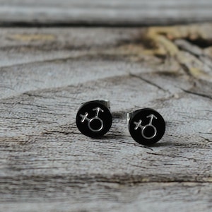 Könnte beinhalten: Ein Paar schwarze Ohrstecker mit einem silbernen Transgender-Symbol auf jedem Ohrstecker.