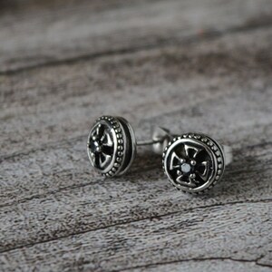 Könnte beinhalten: Ein Paar silberne Ohrstecker mit einem schwarzen Kreuzdesign. Die Ohrringe sind klein und rund und mit einem einzigen schwarzen Stein in der Mitte des Kreuzes besetzt.