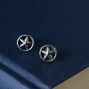 Könnte beinhalten: Ein Paar silberne Stern-Ohrstecker. Die Ohrringe sind rund und haben einen fünfzackigen Stern in der Mitte.