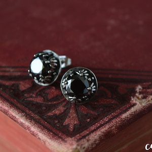 Könnte beinhalten: Ein Paar Ohrstecker mit einem dunklen, facettierten Mittelstein. Die Ohrringe haben eine silberfarbene, floral gemusterte Lünettenfassung. Die Ohrringe werden auf einer dunkelroten, strukturierten Oberfläche präsentiert.