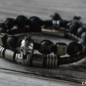 May include: Two stacked bracelets. The top bracelet has black beads and a braided black cord. The bottom bracelet features a brown leather band and silver-toned metal accents, including a Spartan helmet charm and square beads.