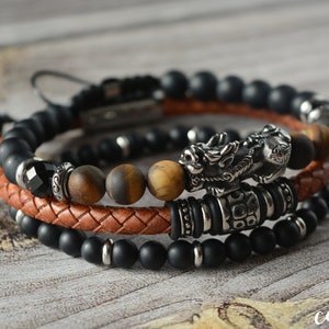 May include: Three layered bracelets with black onyx beads, tiger's eye beads, and a brown leather cord. The middle layer features a silver dragon charm.