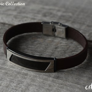 Könnte beinhalten: Ein braunes Lederarmband mit einem silbernen Metallverschluss und einer schwarzen rechteckigen Platte.