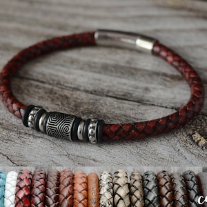 May include: A braided leather bracelet in a deep red colour, with a geometric silver accent and a magnetic clasp. The bracelet is displayed on a wooden surface, showcasing its texture and design. Additional colour options are shown below.