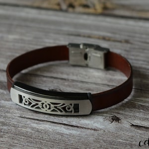 Könnte beinhalten: Ein braunes Lederarmband mit einem silbernen Metallverschluss und einer dekorativen silbernen Metallplatte mit einem Ausschnittmuster.