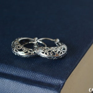 May include: Two silver hoop earrings with a detailed filigree pattern. The earrings are displayed on a dark blue surface, highlighting the delicate openwork design. A classic piece of jewellery.
