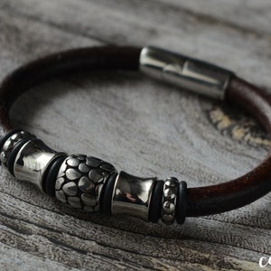 May include: A brown leather bracelet with silver metal detailing. The bracelet has a magnetic clasp and decorative metal beads. The central bead has a textured, pebble-like design. The bracelet is displayed on a wooden surface.