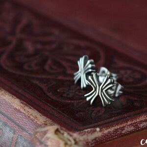 Könnte beinhalten: Zwei silberne Ohrringe in Kreuzform auf einer roten, gemusterten Oberfläche.