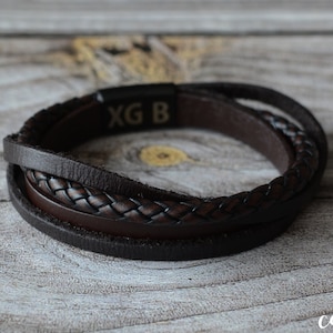May include: A brown leather bracelet with multiple strands and a braided central section. The bracelet features a magnetic clasp with the engraved letters "XG B". It is presented on a wooden surface.
