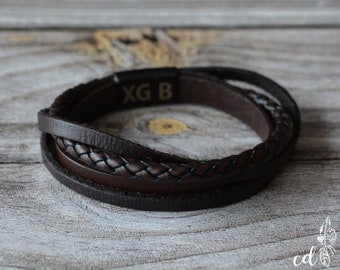 May include: A multi-strand brown leather bracelet with a braided center band. The bracelet has a magnetic clasp with the engraved letters "XG B". The bracelet is displayed on a wooden surface.