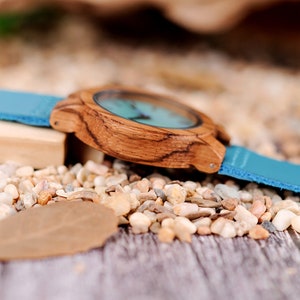 Puede incluir: Un reloj de pulsera de madera con una correa de cuero azul. La esfera del reloj es de color azul claro y la caja del reloj es de madera oscura.