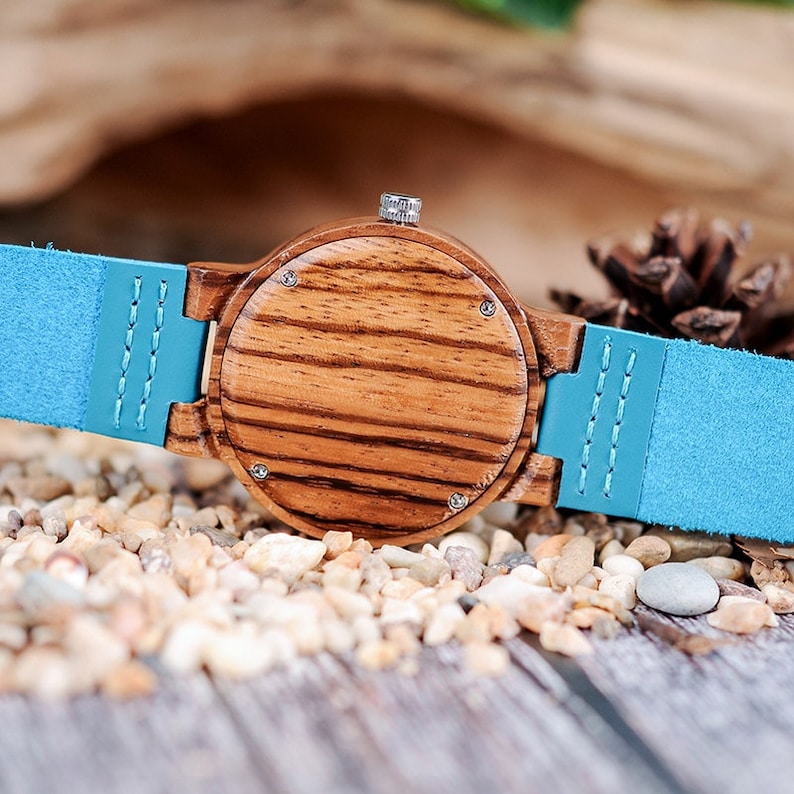 Puede incluir: Un reloj de pulsera de madera con una correa de cuero azul. La parte posterior del reloj est&aacute; hecha de madera con un patr&oacute;n de rayas.