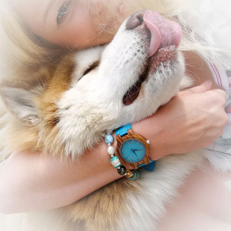Puede incluir: Una mujer lleva un reloj de madera azul y marr&oacute;n con una esfera azul y un brazalete de cuentas. La mujer est&aacute; sosteniendo un perro con la lengua fuera.