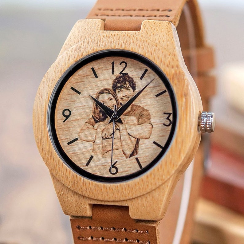 May include: A wooden wristwatch with a brown leather band. The watch face has a black background with white Roman numeral hour markers and a black and white photo of a couple.