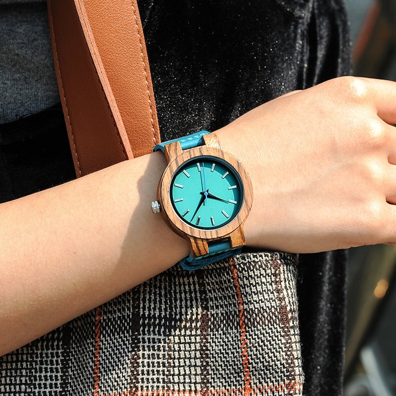 Puede incluir: Un reloj de pulsera de madera con una esfera azul y una correa de madera marr&oacute;n. El reloj est&aacute; en la mu&ntilde;eca de una persona.