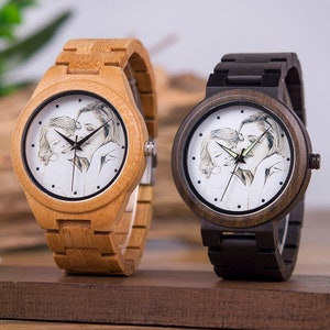 May include: Two wooden wristwatches with white faces featuring a black and white photo of a mother and child. The watch on the left has a light wood band and the watch on the right has a dark wood band.