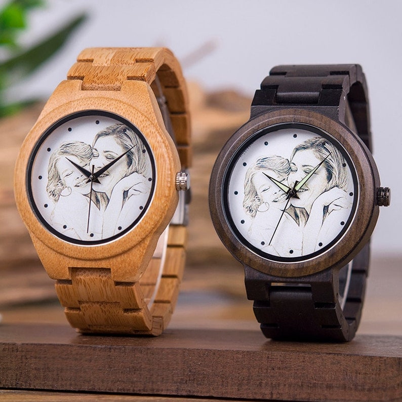 May include: Two wooden wristwatches with white faces featuring a black and white photo of a mother and child. The watch on the left has a light wood band and the watch on the right has a dark wood band.