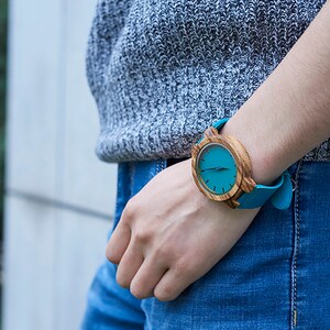 Puede incluir: Un reloj de pulsera de madera con una esfera azul turquesa y una correa de cuero azul turquesa.