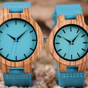 Puede incluir: Dos relojes de pulsera de madera con correas de cuero azul y esferas de color azul claro. Los relojes tienen agujas de hora y minuto negras y marcadores de hora negros.