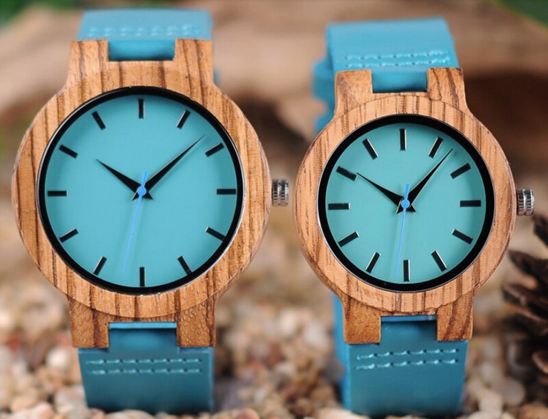 Puede incluir: Dos relojes de pulsera de madera con correas de cuero azul y esferas de color azul claro. Los relojes tienen agujas de hora y minuto negras y marcadores de hora negros.