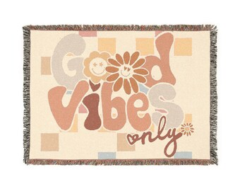 May include: A woven throw blanket with a cream background and a colorful design that says "Good Vibes Only" in a retro font. The design includes flowers and smiley faces.