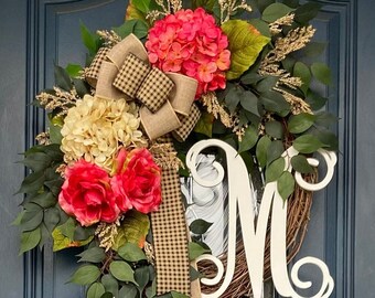Può includere: Una ghirlanda decorativa con base intrecciata, adornata con vibranti fiori rosa e crema, lussureggiante fogliame verde e un grande fiocco color cammello con un motivo a scacchi bianco e nero. Una lettera bianca "M" è attaccata alla ghirlanda.
