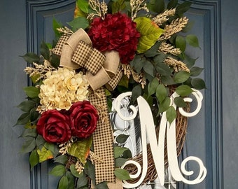 Può includere: Una ghirlanda con una grande lettera bianca "M" al centro. La ghirlanda è decorata con fiori bordeaux e crema, verde e un nastro a quadri marrone e bianco.