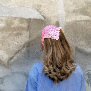 May include: A pink sequined headpiece with a scalloped edge. The headpiece is worn over long, wavy brown hair. The person is wearing a blue and white striped button-down shirt.