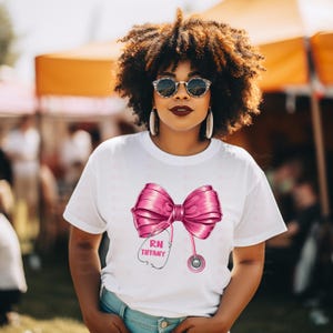 May include: White t-shirt featuring a large pink bow design with a stethoscope and the text "RN Tiffany". The bow is detailed with shading, and the stethoscope has a pink head. The shirt is a casual style.