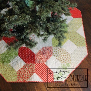 Pode incluir: Uma saia de árvore de Natal em patchwork vermelho, verde e branco com uma borda festonada. A saia é acolchoada e apresenta uma variedade de padrões florais e geométricos. O texto "The Tipsy Needle by Sew Chic Fabric" está impresso na saia.