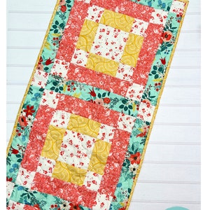 Peut inclure: Un modèle de courtepointe pour chemin de table avec un motif floral dans les tons de jaune, rouge, blanc et vert. Le motif s'appelle "All Boxed Up Table Runner" et mesure 16 pouces par 72 pouces. Le modèle est de The Tipsy Needle et est décrit comme rapide et facile, idéal pour les débutants.