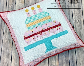 May include: A white pillow with a red border and a pink and red inner border features a quilted birthday cake design. The cake is made of various patterned fabrics in pink, red, yellow, blue, and green. The cake has three yellow candles and is on a blue stand. The pillow has the text "The Tipsy Needle By Sew Chic Fabrics" in the top right corner.