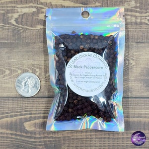 May include: A clear, iridescent bag filled with whole black peppercorns. The bag has a circular label with the text "Black Peppercorn" and "Witchmade Studios". A quarter is next to the bag. The net weight is 1 oz (28.35 grams).