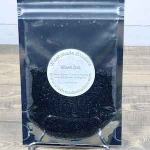 May include: A small clear plastic bag filled with black salt. The bag has a white label with the text "Witchmade Studios" and "Black Salt" in black lettering. The label also includes the text "Removing jinxes. Protection. Keeping Away Bad Elements or Neighbors" and "1 oz net wt 28.35 gm". The website address "www.witchmadestudios.com" is printed on the label in a circle.