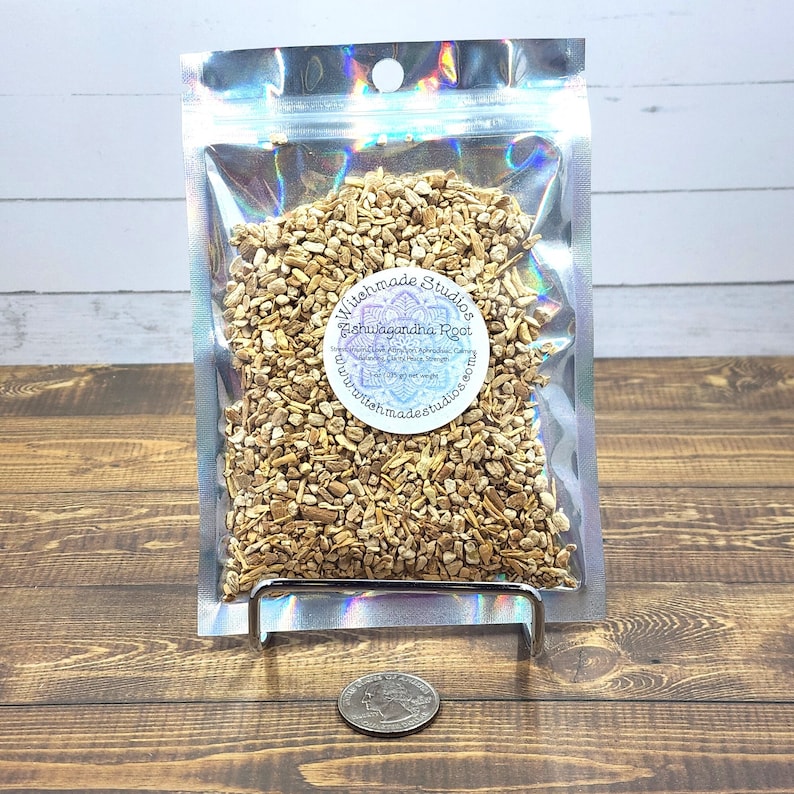 Pode incluir: Um saco de fecho herm&eacute;tico prateado cheio de peda&ccedil;os de raiz de Ashwagandha seca de cor castanho claro. O saco tem um r&oacute;tulo branco com o texto "Witchmade Studios Ashwagandha Root" e um endere&ccedil;o de site.