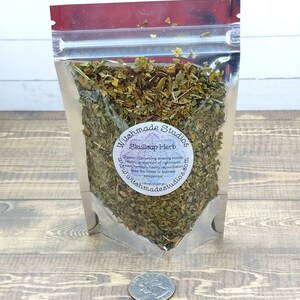 May include: A clear plastic bag filled with dried green herbs labeled "Skullcap Herb" with the text "Witchmade Studios" and a website address. The bag is sitting on a wooden surface.