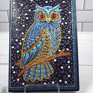 May include: A black journal cover adorned with a detailed owl design. The owl is rendered in shades of blue, gold, and orange, with red eyes, and is embellished with sparkling crystals. The background is a dark blue, speckled with white and purple accents.