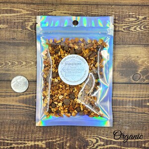 May include: A clear, iridescent pouch filled with a mix of brown spices, including whole peppercorns. The pouch has a circular label that reads "Witchmade Studios Mulling Spices". The word "Organic" is printed in cursive in the bottom right corner.