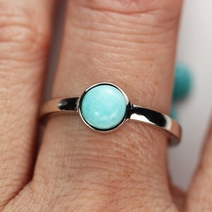 May include: A silver ring featuring a round, light blue gemstone. The gemstone is set in a simple, polished silver band. The ring is shown on a finger, with a blurred background.