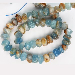 May include: A strand of faceted, irregular gemstone beads in shades of light blue and translucent white with streaks of warm brown. The beads are strung on a thin blue thread, displaying a polished, multi-faceted surface texture throughout.